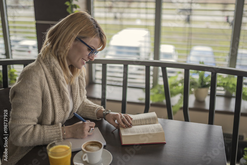 Young smiling woman in a restaurant reading a book and taking notes while drinking a coffee