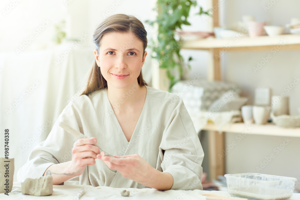 Pretty young woman looking at camera by her workplace while making kitchenware from white clay Pretty young woman looking at camera by her workplace while making kitchenware from white clay