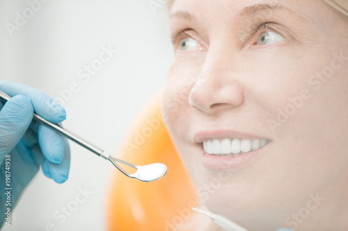 Healthy mature woman with toothy smile looking at dentist with mirror before check-up