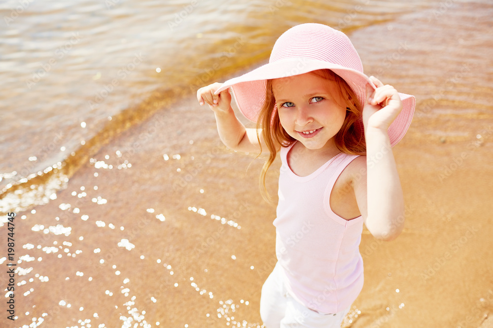 Cute little girl in elegant hat looking at camera while spending summer vacation by waterside