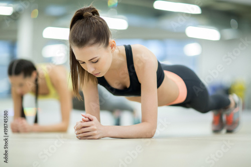 Young woman in activewear doing planks on the floor of gym with another girl on background