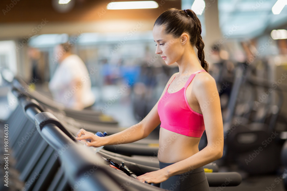 Fit girl in activewear choosing run speed on display while going to work out on treadmill in gym Fit girl in activewear choosing run speed on display while going to work out on treadmill in gym