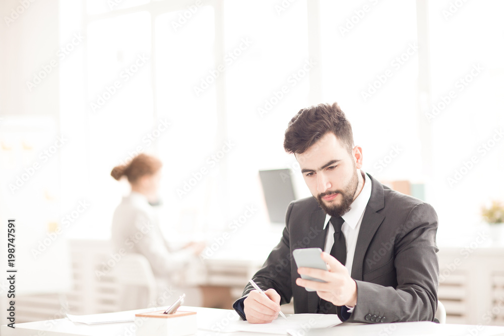 Young businessman using his phone and making notes at the table at office Young businessman using his phone and making notes at the table at office