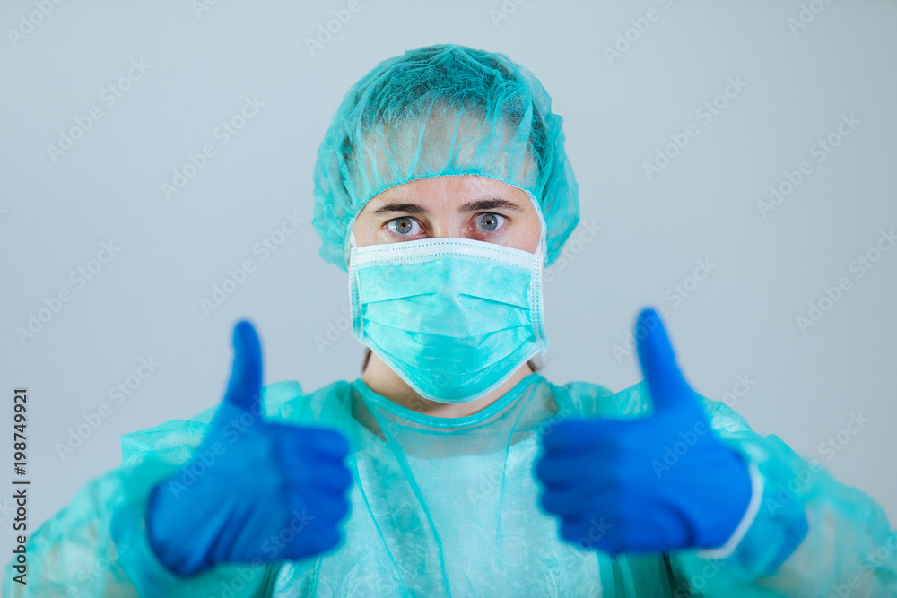 Young woman doctor with blue eyes wearing a cap and face mask show sight thumbs up and good luck Young woman doctor with blue eyes wearing a cap and face mask show sight thumbs up and good luck