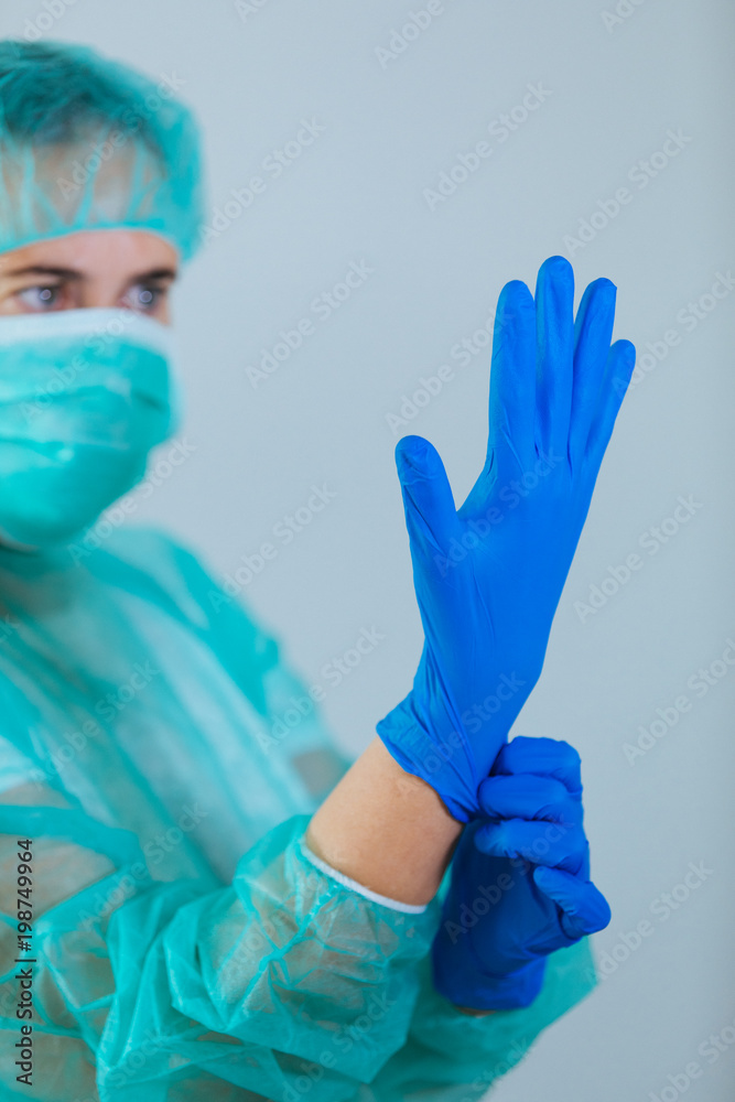 Fototapeta premium Young woman doctor with blue eyes wearing a cap and face mask putting gloves