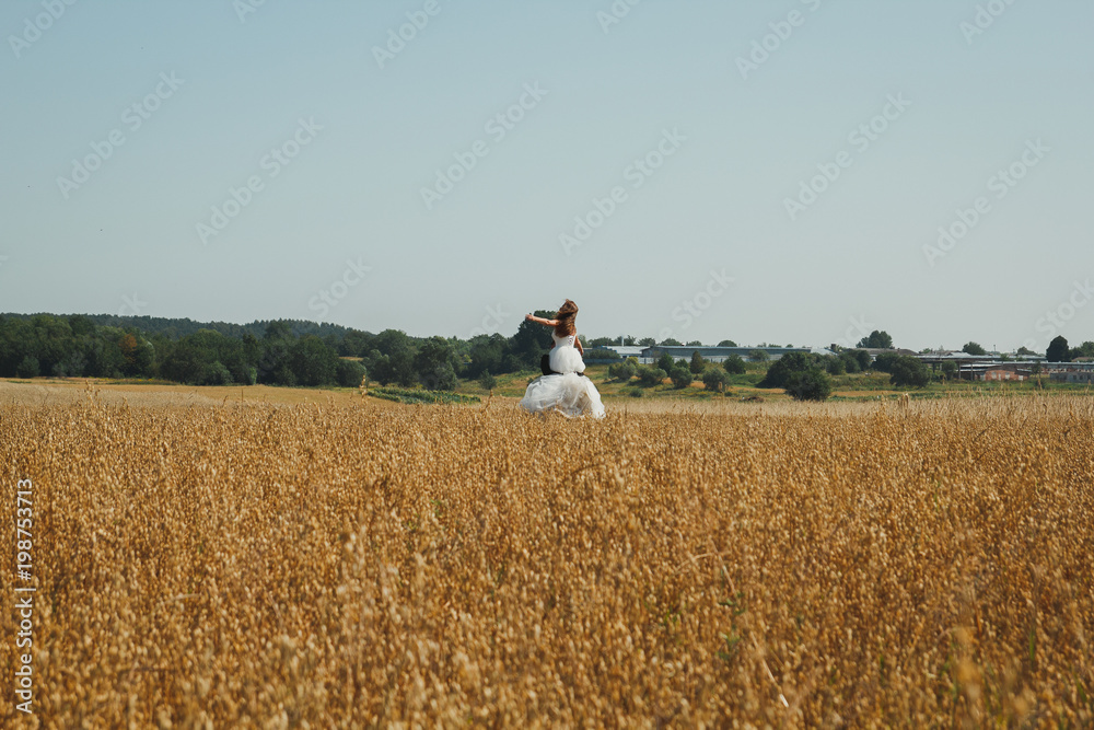 Obraz premium Wedding couple is spinning in the summer wheat field. Bride in white puffy dress hold groom's hands in black suit. Green trees and houses on the horizon. Summer heat and love story.