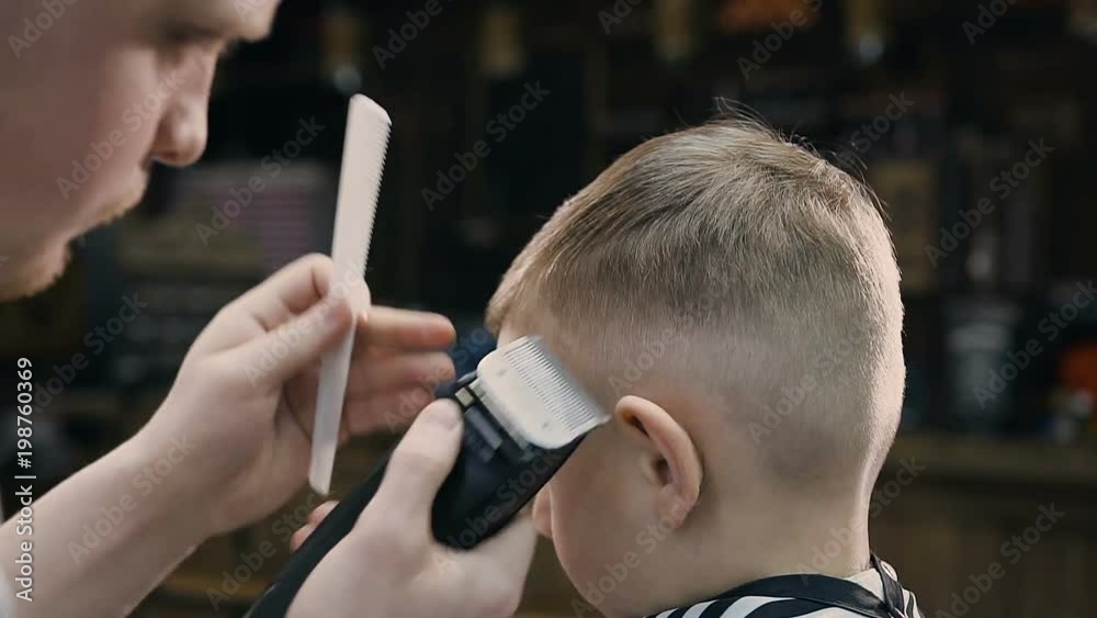 Close-up. Hairdresser with cute client child in the barbershop. Barber using trimmer and comb in barbershop. Beautiful cute kid sitting on the chair in hairdressing salon Close-up. Hairdresser with cute client child in the barbershop. Barber using trimmer and comb in barbershop. Beautiful cute kid sitting on the chair in hairdressing salon