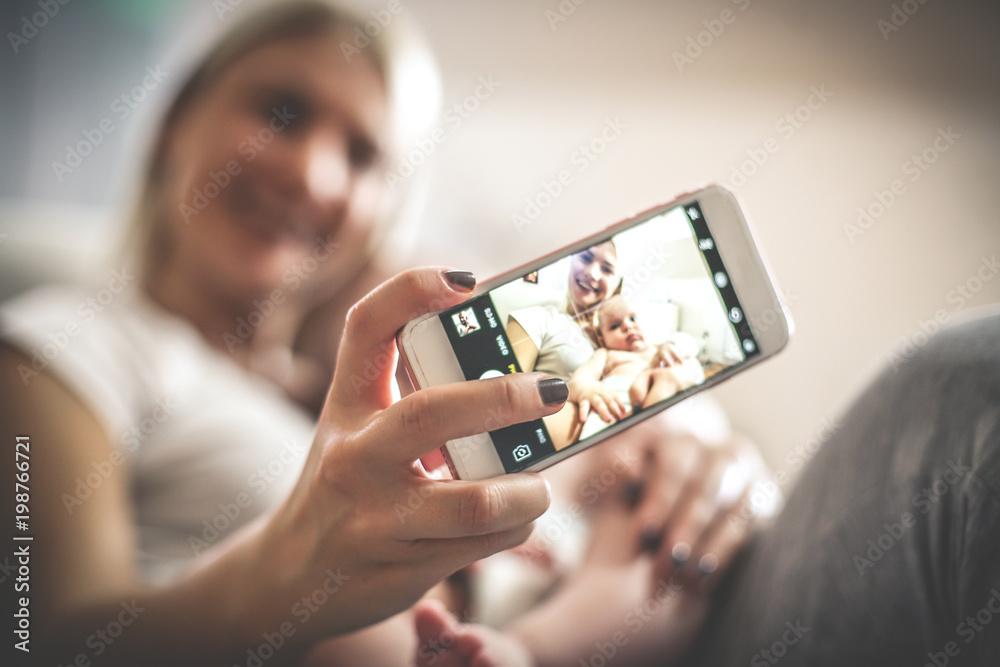 Smiley mother with her baby boy at home taking self portrait with mobile phone. Focus is on hand.