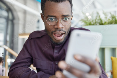 Shocked dark skinned businessman in eyewear, recieves notification on modern smart phone, gets notice to pay bills, has surprised expression. African American male reads shocking news in internet.