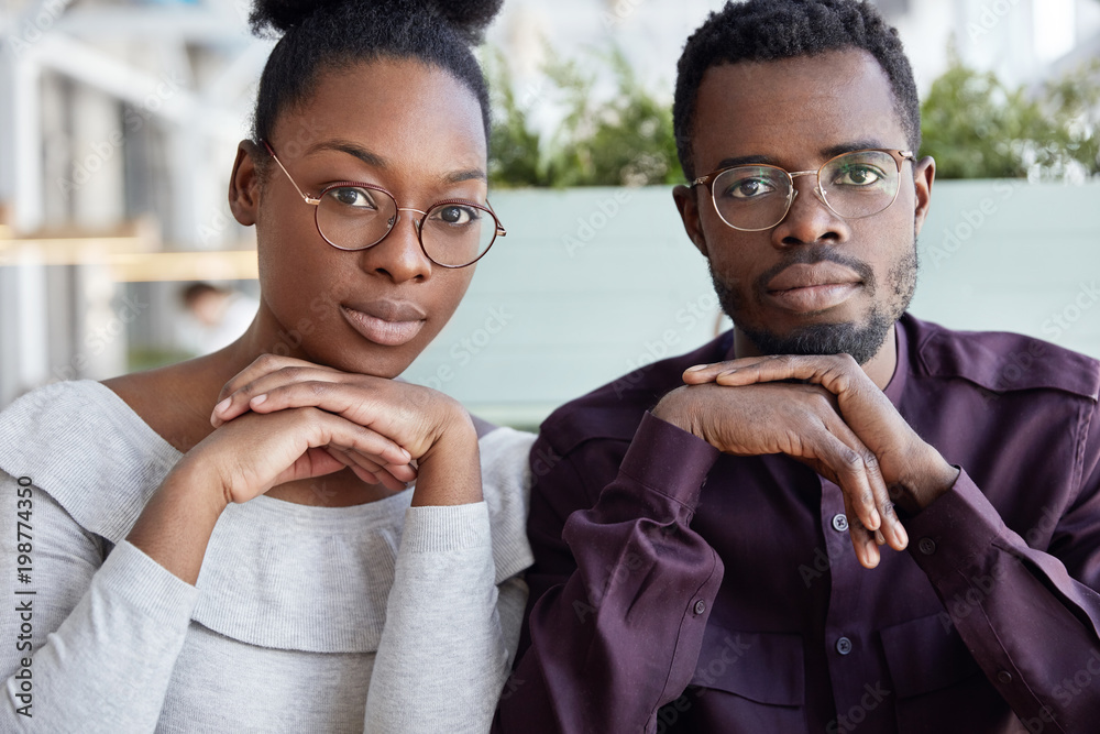 Fototapeta premium Teamwork and cooperation concept. Successful African American female and male colleagues sit close to each other, wears spectacles, work on common project. Business partners have meeting in cafe