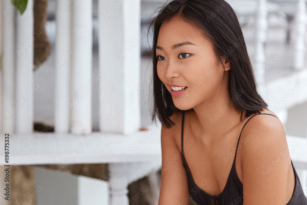 Sideways shot of beautiful glad mixed race female with dark hair, focused on something with cheerful expression, thinks about something pleasant, plans coming weekend or holidays. Chinese woman indoor Sideways shot of beautiful glad mixed race female with dark hair, focused on something with cheerful expression, thinks about something pleasant, plans coming weekend or holidays. Chinese woman indoor