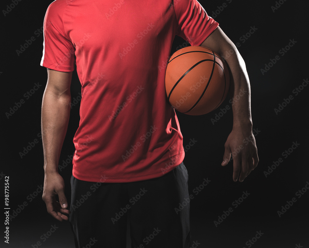 Young athletic man in red t-shirt with orange ball in armpit. Isolated on black background. Close up of his body Young athletic man in red t-shirt with orange ball in armpit. Isolated on black background. Close up of his body