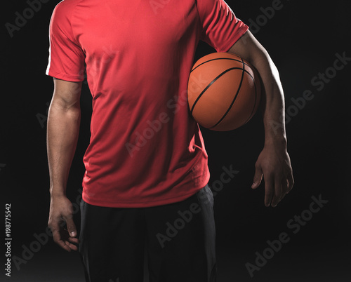 Young athletic man in red t-shirt with orange ball in armpit. Isolated on black background. Close up of his body