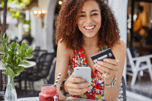 Internet banking and ecommerce concept. Happy young smiling female with Afro hairstyle, uses modern cell phone and credit card for online shopping, enjoys fresh fruit smoothie in terrace bar. photo