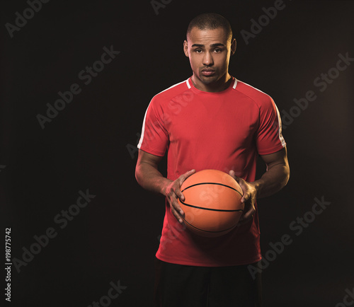 Should play ball or not. Waist up portrait of handsome african guy in red t-shirt wth basketball. He watching at camera with confused look. Isolated on black background