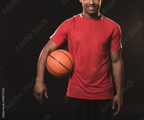 Young athletic sportsman keeping basketball with right armpit and smiling. Isolated on black background