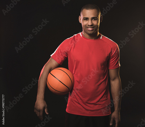 Waist up portrait of handsome african sportsman. Guy holding the basketball with right armpit while happily smiling. Isolated on black background