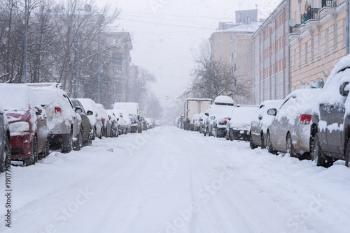 Snowfall in the empty street. Rows of parked cars covered with snow. Buildings and park can be seen through spots of falling snowflakes. White winter in a city