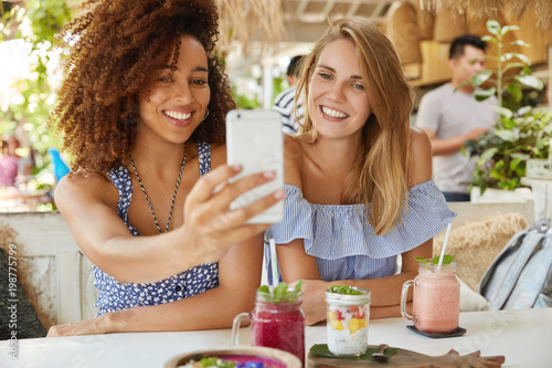 Photo of glad mixed race females have interracial friendship, pose at camera of modern cell phone, make selfie while have rest in cozy terrace bar, enjoy fresh drinks. People, ethnicity and leisure