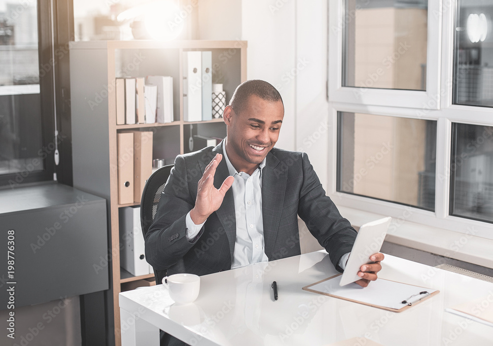 Young handsome guy sitting at office and communicating on video using  tablet. Man waving hand while looking at screen with happily smile Stock  Photo | Adobe Stock