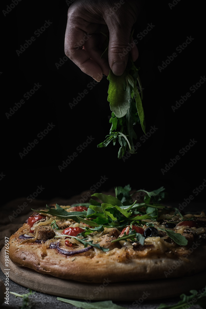 Fresh pizza with tomatoes, cheese, chicken and mushrooms. Italian pizza sprinkled arugula, tired of labor by hands. Black rustic background. Closeup