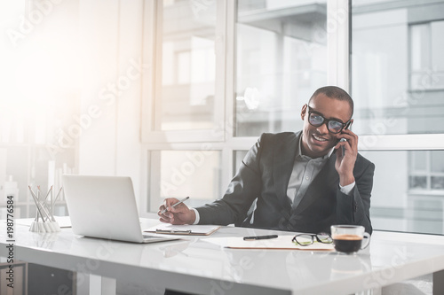 Portrait of young african man sitting at office and talking on phone. He with happy smile is looking at camera. Guy ready making notes in document. Copy space in left side