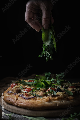 Obraz na płótnie Fresh pizza with tomatoes, cheese, chicken and mushrooms. Italian pizza sprinkled arugula, tired of labor by hands. Black rustic background. Closeup w salonie