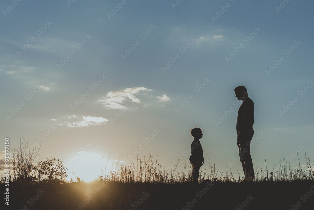 Side view profile of adult and little male silhouette standing on grass opposite each other against blue sky background. Copy space in left side
