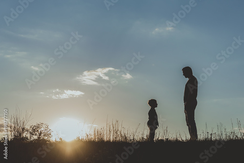 Side view profile of adult and little male silhouette standing on grass opposite each other against blue sky background. Copy space in left side