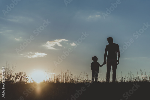 Silhouette of father and son standing on grass and holding hands against blue sky background. Copy space in left side