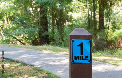 A one mile marker sign post beside a paved pathway with a bright green and sunny background.