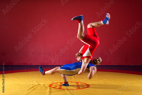 Two young men in blue and red wrestling tights are wrestlng and making a suplex wrestling on a yellow wrestling carpet in the gym