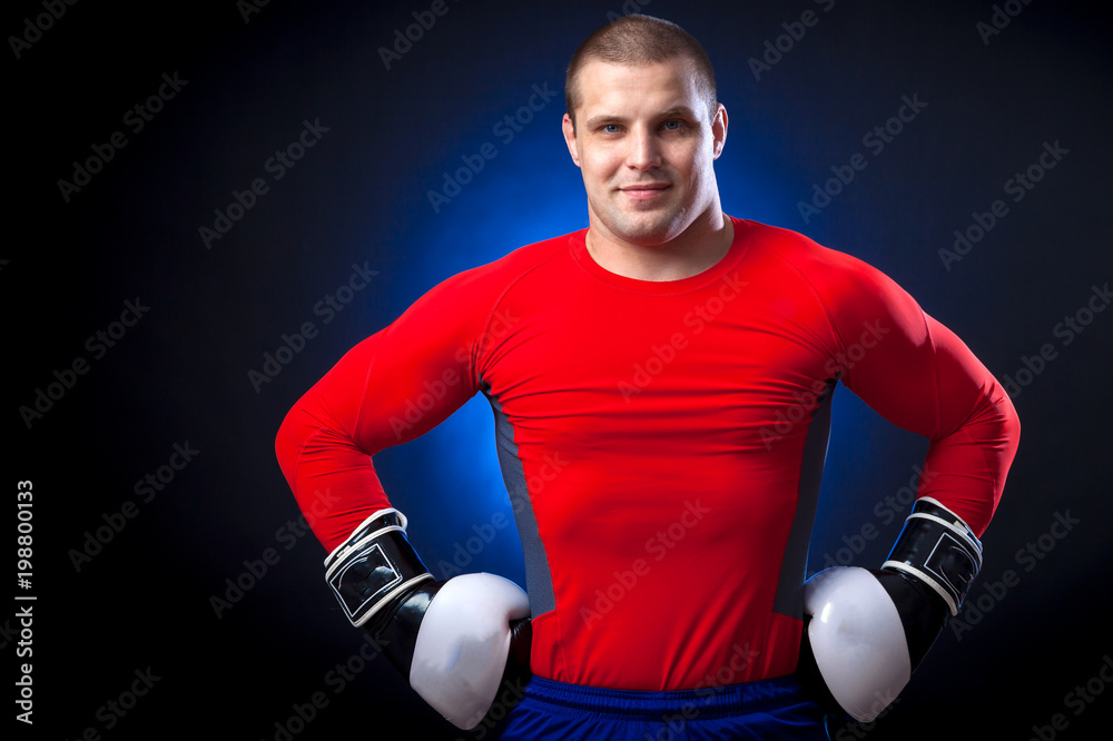 A strong dark-haired sportman boxer in a red sports wear  rush guard and  in black and white boxing gloves smile and standing against a blue a lights on a black isolated  background