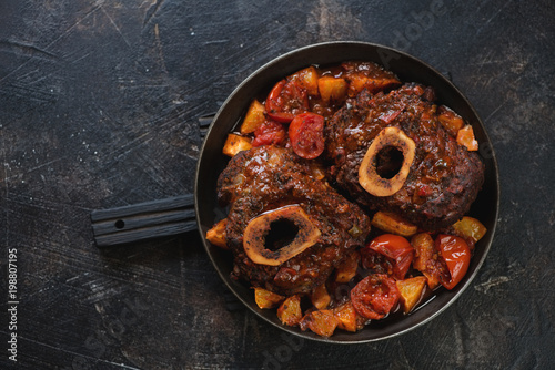 Metal pan with ossobuco made of cross cut veal shank with oranges and tomatoes. Above view on a dark brown stone background, horizontal shot with space