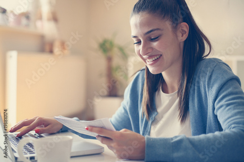 Cheerful young woman at home reading bills and typing on laptop.