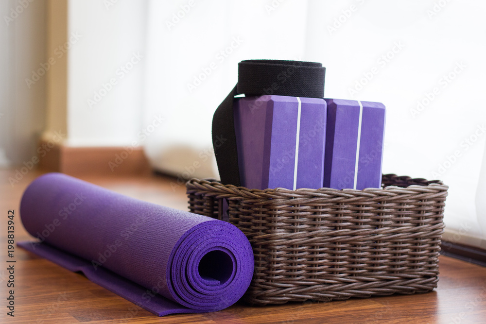 Purple yoga mat rolled by wicker basket with blocks and black belt inside.  Yoga props on center wooden floor by window white curtain. Healthy  lifestyle activity concept Stock Photo