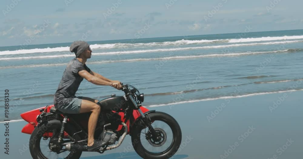 Handsome man biker surfer driving his black motorbike cafe racer with red surfboard shortboard on the beach along the ocean at sunny day. 4k video shooting by handheld gimbal Handsome man biker surfer driving his black motorbike cafe racer with red surfboard shortboard on the beach along the ocean at sunny day. 4k video shooting by handheld gimbal