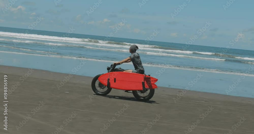 Handsome man biker surfer driving his black motorbike cafe racer with red surfboard shortboard on the beach along the ocean at sunny day. 4k video shooting by handheld gimbal Handsome man biker surfer driving his black motorbike cafe racer with red surfboard shortboard on the beach along the ocean at sunny day. 4k video shooting by handheld gimbal