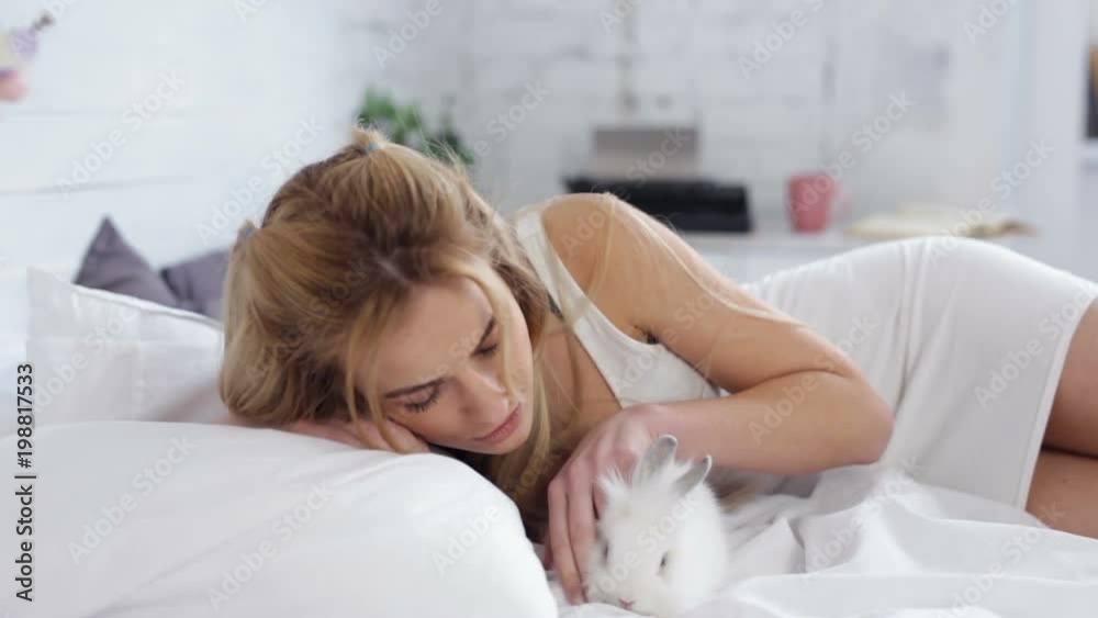 Pretty blonde woman hugging and kissing white rabbit and looking at camera. She lies in bed at the morning. Pretty blonde woman hugging and kissing white rabbit and looking at camera. She lies in bed at the morning.