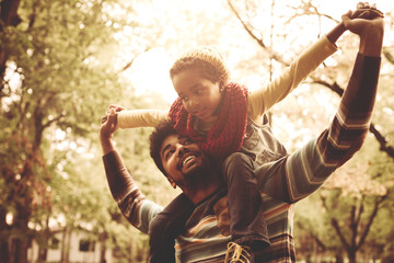 Cheerful African American little girl sitting on father shoulders in park.