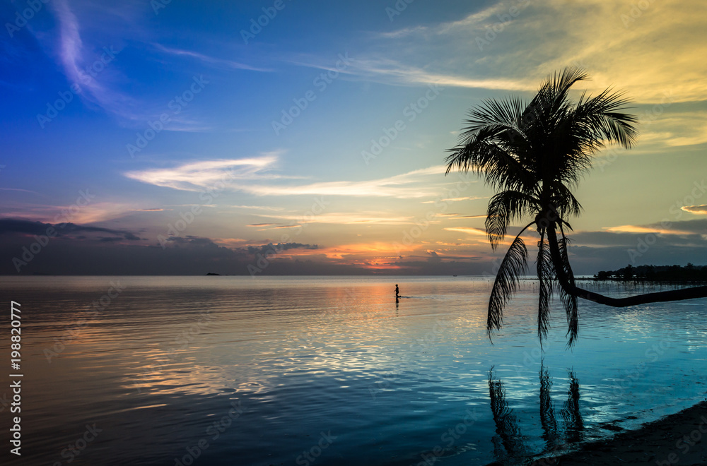 Peaceful blueish sunset landscape on the beach with silhouette of palm tree and man walking on shallow water in the island of Koh Phangan, Thailand