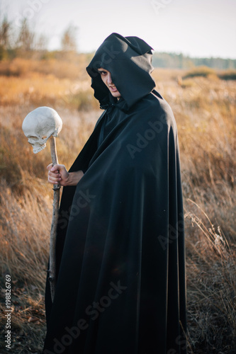 Halloween image. A handsome guy in a black cloak with a staff from a human skull against the background of a yellow field. Fantasy photo