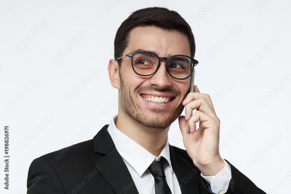 Closeup shot of young business man isolated on grey background wearing stylish eyeglasses enjoying telephone conversation with happy toothy smile, laughing at joke Closeup shot of young business man isolated on grey background wearing stylish eyeglasses enjoying telephone conversation with happy toothy smile, laughing at joke