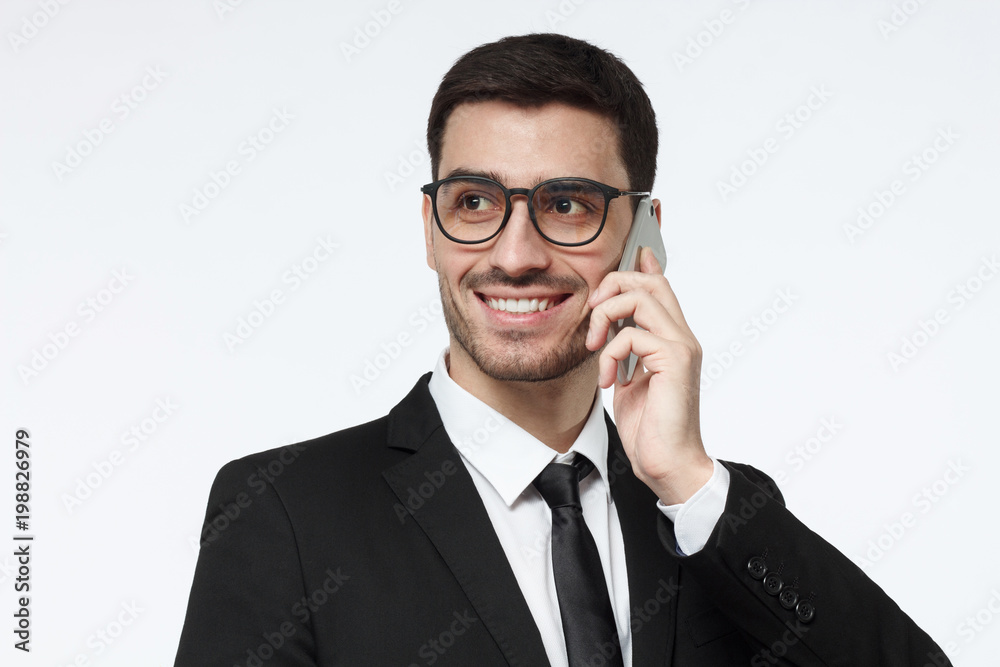 Closeup picture of young smiling business man pictured isolated on grey background with trendy eyeglasses on, pressing phone to ear while having conversation with colleague Closeup picture of young smiling business man pictured isolated on grey background with trendy eyeglasses on, pressing phone to ear while having conversation with colleague