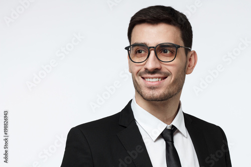 Closeup shot of young corporate advisor in trendy spectacles pictured isolated on grey background wearing formal black suit, showing toothy open smile, looking to the left, positive and confident