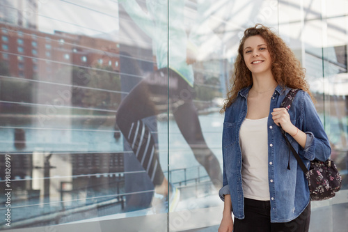 Adorable lovely glad young female with curly hair, wears fashionable denim jacket and carries rucksack, comes in shopping centre for buying new clothes and products, enjoys spare time or weekend photo