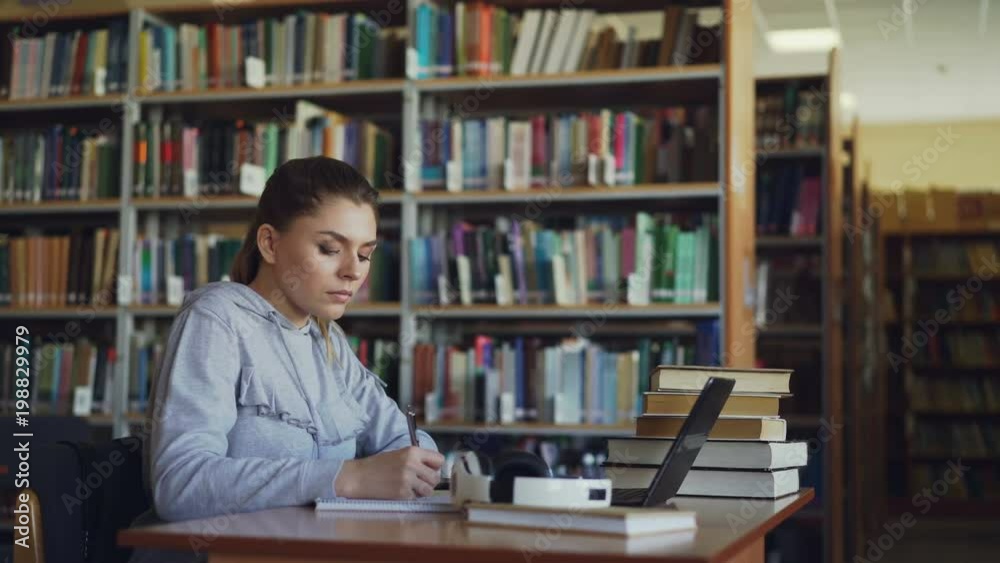 Beautiful serious caucasian female student sitting at table with books in library writing down summary in copybook using laptop computer Beautiful serious caucasian female student sitting at table with books in library writing down summary in copybook using laptop computer