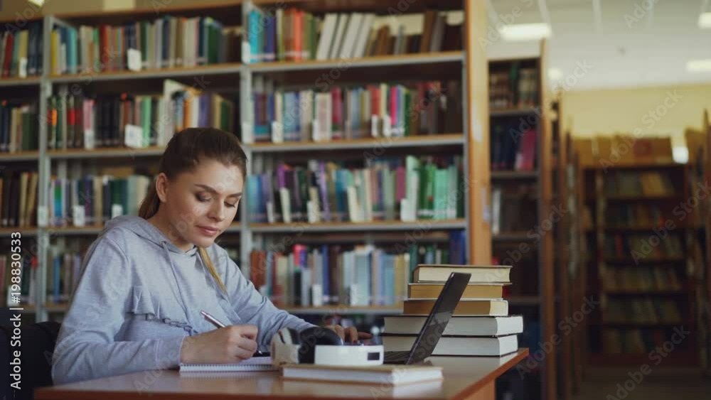 Young beautiful smiling student girl is sitting at desk in big spacious library with pile of books. She is writing something in copybook, laptop is in front of her Young beautiful smiling student girl is sitting at desk in big spacious library with pile of books. She is writing something in copybook, laptop is in front of her