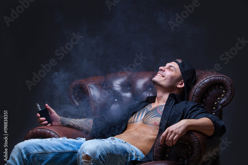 Vaper. The man with tattoos sits on a leather sofa smoke an electronic cigarette on the dark background. photo