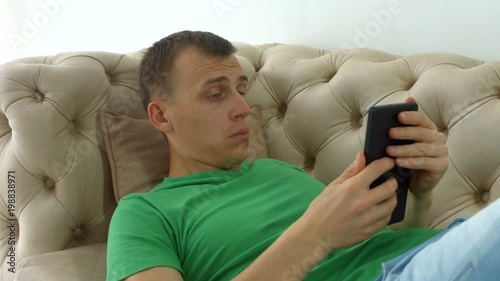 Positive handsome man networking on digital tablet while lying on sofa in domestic room. Amuzed male hipster browsing social media content on tablet computer while spending leisure at home. Closeup. photo